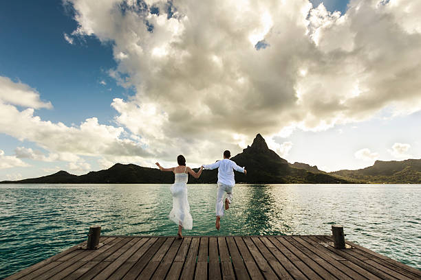 Young Couple Celebrating a Polynesian Wedding in Bora Bora A DSLR photo of a newlywed couple jumping from a wood pier into the wonderful sea of Bora Bora, French Polynesia. It is just after sunset, so all scene is very soft lit. She is wearing a white wedding dress. In the background, silhouetted against the sky, is mountain Otemanu, main natural landmark in Bora Bora. polynesia photos stock pictures, royalty-free photos & images