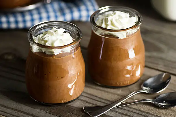 Homemade chocolate pudding topped with whipped cream in little glass cups on a wooden table. Two spoons are in the foreground.