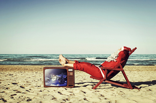 Santa Claus on the beach, relaxing and sunbathing with feet resting on a vintage TV.