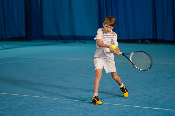 Young Tennis Player Practicing Indoors Determined young boy old keeps his eye on the tennis ball as he swings. backhand stroke stock pictures, royalty-free photos & images