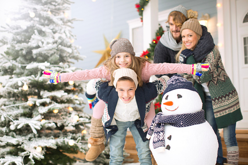 Family with two kids having fun outdoors in front of house, playing with snowman. Brother carrying his sister ob his back. Wearing knitted sweaters, hats and scarfs. House, yard and tree are decorated with festive string lights. Fir tree in background covered with snow. Day time.