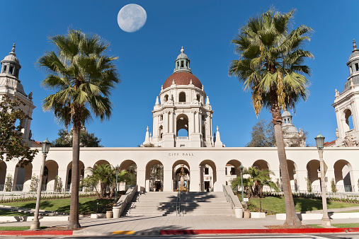 A picture of the Pasadena City Hall in California, USA. The image was taken looking east on a morning with clear blue sky. A telephoto shot a the setting moon was taken separately, and a digital composite was created. There are two beautiful palm trees symmetrically planted relative to the building. Most of the west arcade is shown. No people are present in this color image.