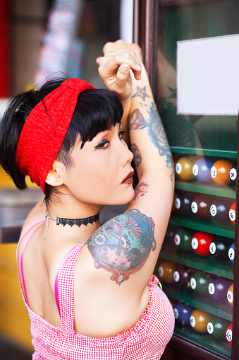 Thai girl woth tattoos in retro fashion leaning at cabinet with pool balls.