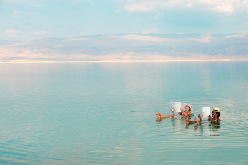 Lovely girls floating in salty water of Dead Sea and reading newspaper. Unusual buoyancy caused by high salinity.