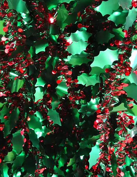 Background photo of sparkling coloured tinsel Christmas decorations / foil garland, with shiny green holly leaves and bunches of red berries.