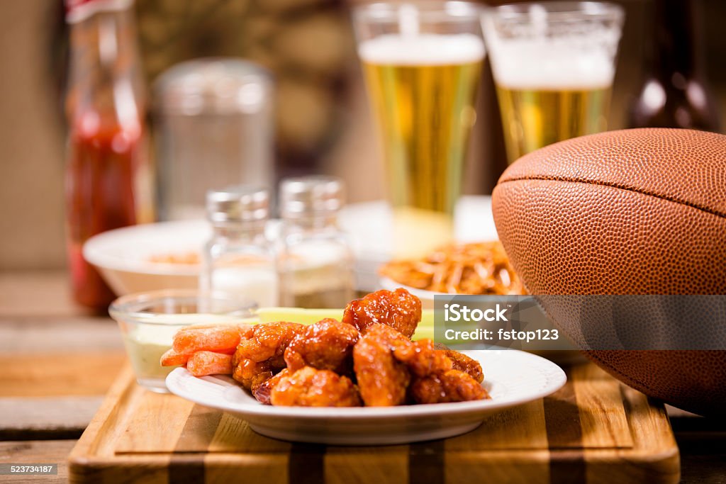 Pub food. Snacks, beer, football. Spicy chicken wings. Sports bar, pub setting. Spicy chicken wings foreground. Football, pretzels, peanuts, beer on bar counter top. Dartboard in background.     American Football - Ball Stock Photo