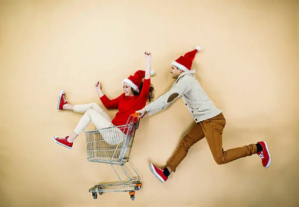 Young couple in Christmas hats having fun running with shopping trolley against the beige background