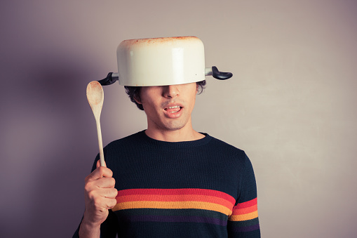 A silly young man is wearing a cooking pot on his had and is holding a wooden spoon