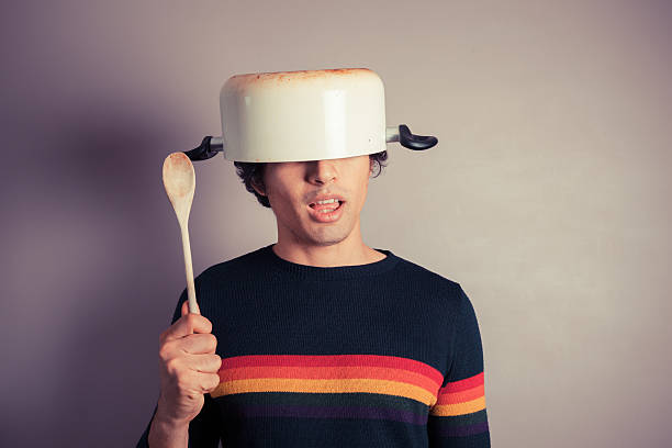 Silly young man with pot on his head A silly young man is wearing a cooking pot on his had and is holding a wooden spoon fool stock pictures, royalty-free photos & images