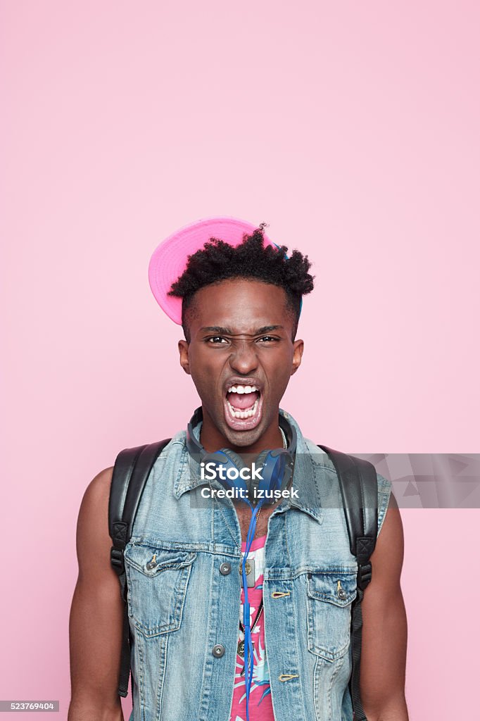 Portrait of angry afro american guy Portrait of angry afro american young man wearing headphone, cap and jeans sleeveless jacket, standing against pink background, shouting. Adult Stock Photo