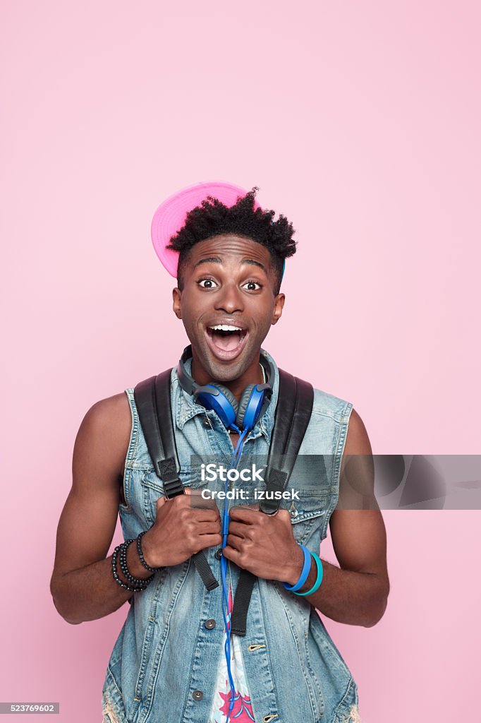 Summer portrait of excited afro american guy Portrait of surprised afro american young man wearing headphone, cap and jeans sleeveless jacket, standing against pink background, rollining eyes, shouting at camera. Colored Background Stock Photo