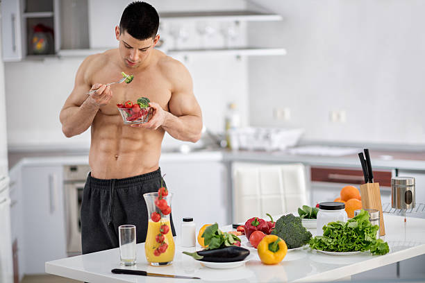 Bodybuilder in the kitchen Young shirtless male fitness athlete standing in the kitchen and holding a bowl with vegetables. There are kitchen knives, orange juice and more vegetables on the table in front of him. eating body building muscular build vegetable stock pictures, royalty-free photos & images