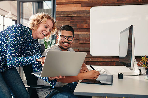 Graphic designers cooperating in work Shot of two happy young graphic designers working in their office with man sitting at his desk and female colleague showing something on her laptop. photo editor photos stock pictures, royalty-free photos & images