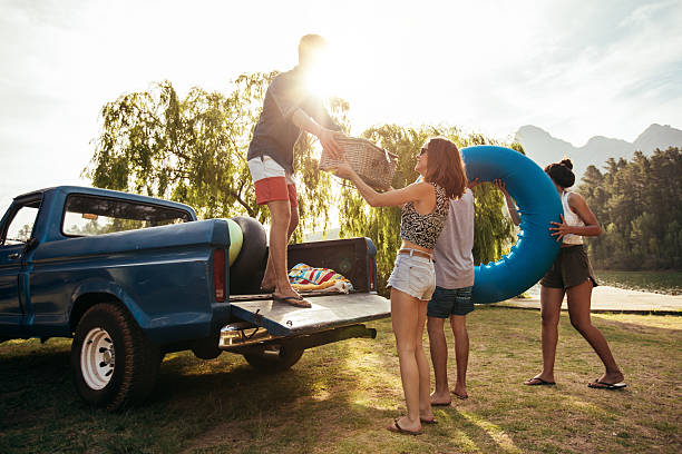 Group of young friends on picnic by the lake. Men and women unloading pickup truck on camping trip, carrying picnic basket and inflatable tube on sunny day.