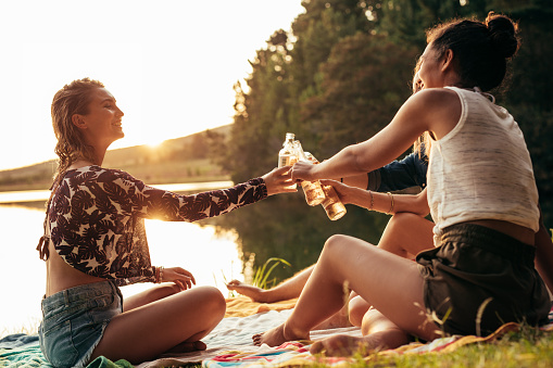Group of young women celebrating at a lake. Young friends are toasting each other with beers during sunset at the lake.