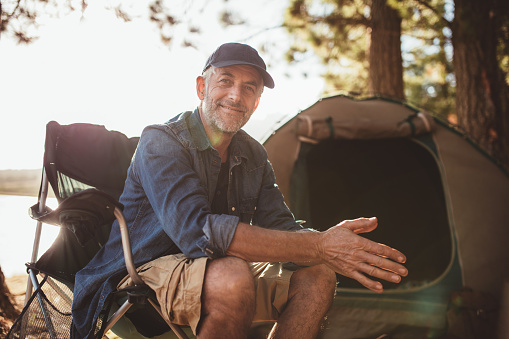 Portrait of a happy mature man sitting in front of a tent and looking at camera. Senior caucasian man at campsite.
