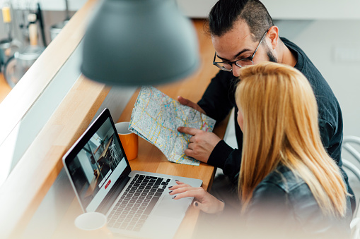 Young Hipster Couple, backpackers, looking on their laptop to rent apartment using vacation home rental services online. Looking at map and laptop. They are cheerful together. Closeup from above.