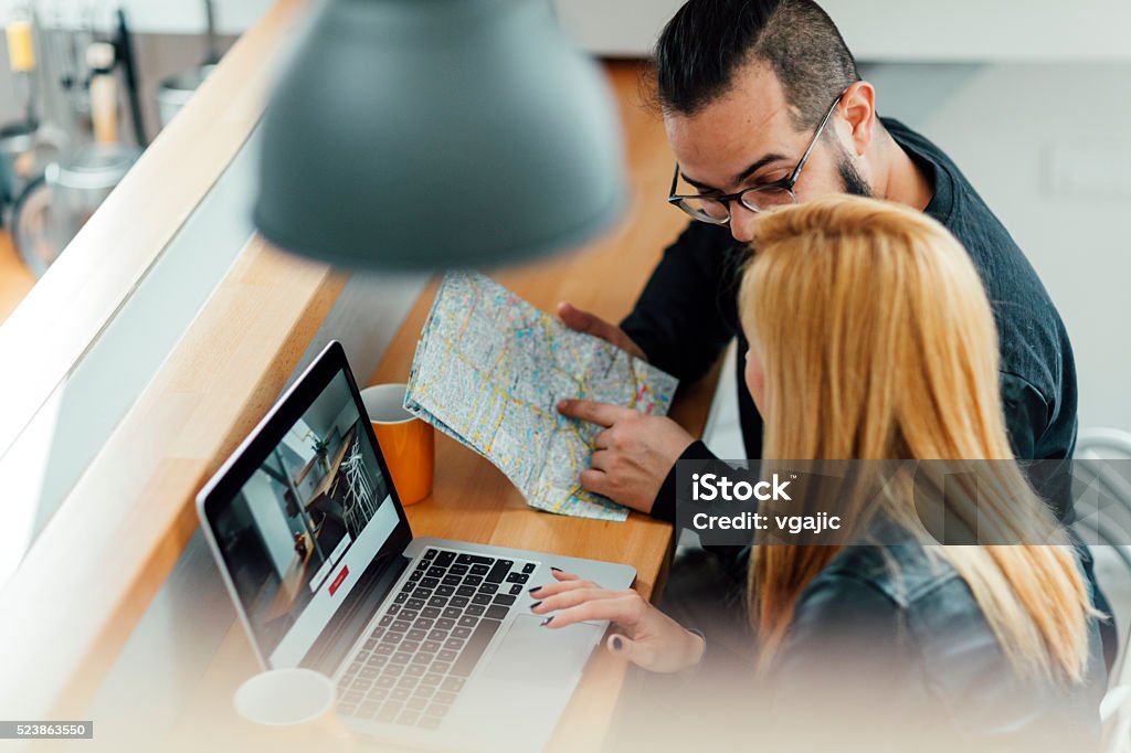 Backpackers Looking For Apartment On Their Laptop. Young Hipster Couple, backpackers, looking on their laptop to rent apartment using vacation home rental services online. Looking at map and laptop. They are cheerful together. Closeup from above. Searching Stock Photo