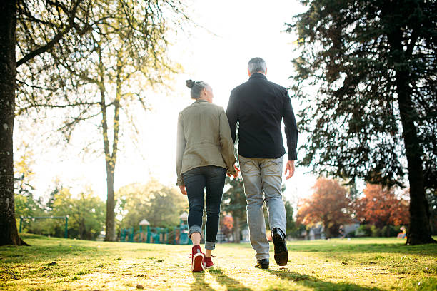 A gray haired and youthful looking couple in their 50's enjoy time with each other in a beautiful outdoor setting, the sun casting a golden glow on the scene.  They hold hands as they walk away from the camera.  Horizontal image with copy space.