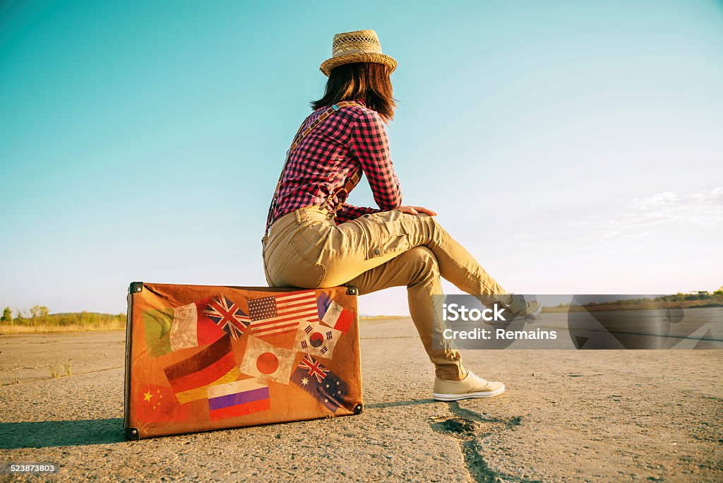 Traveler sits on suitcase with stamps flags Traveler woman sits on retro suitcase and looks away on road. Suitcase with stamps flags representing each country traveled. Suitcase Stock Photo