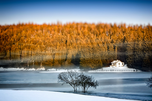 Horizontal composition photography of a large frozen lake in winter season, with a small white cabin in middle of forest with orange sunlight from twilight and some fog / mist due to humidity.