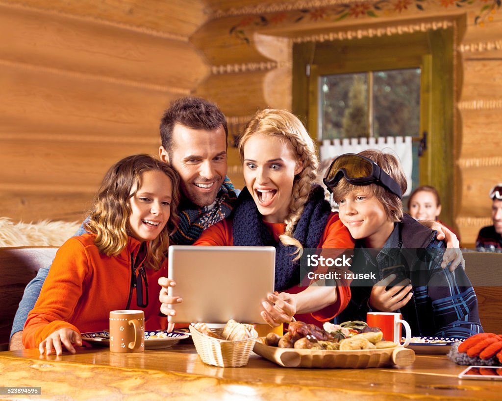 Family having lunch after skiing, using digital tablet Excited Mother, father and their children wearing winter clothes sitting in a mountain restaurant after skiing, having lunch and using a digital tablet. Adolescence Stock Photo