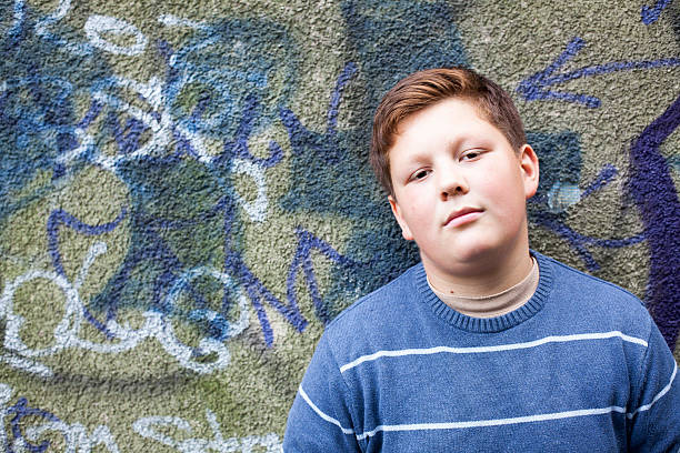 teenage boy portrait in front of graffiti wall overweigh teenage boy with a distanced view leans against a wall with graffiti overweight boy stock pictures, royalty-free photos & images