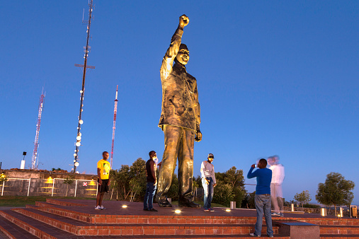 Bloemfontein, South Africa - November, 14th 2014: Teenagers posing for pictures with Nelson Mandela statue that overlooks down on Bloemfontein, situated on Naval Hill. Telecommunication aerials can be seen behind, on a sunset sky.