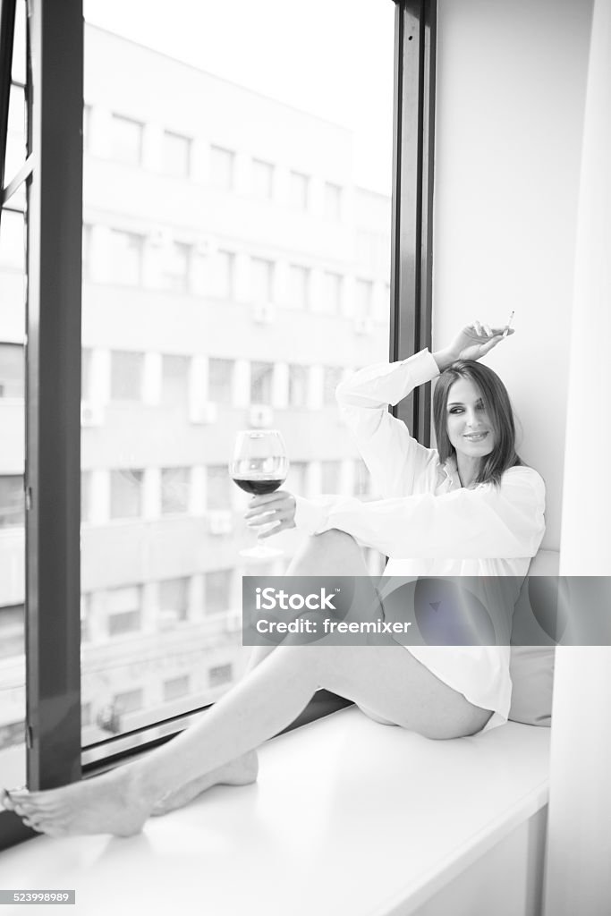 Happy woman drinking wine sitting alone by the window Happy woman drinking wine, smoke cigarette and sitting alone by the window Adult Stock Photo