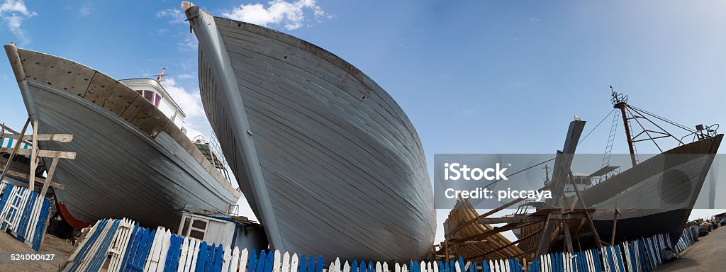 Wooden fishing boats under construction in shipyard, Morocco Panorama of wooden fishing boats under construction in shipyard in the harbour of Essaouira against a blue clear sky, Morocco. Blue Stock Photo