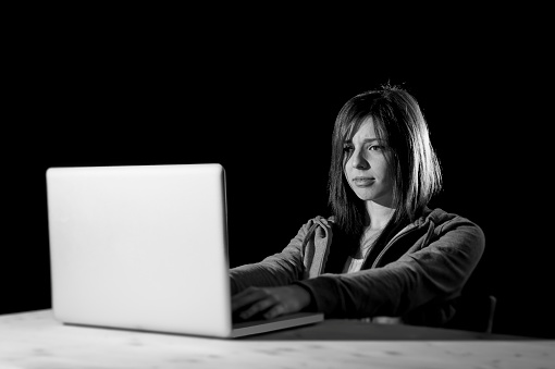 young attractive teen woman looking dark and dangerous hacking laptop computer system on black background in cybercrime or cyber crime and internet criminal concept