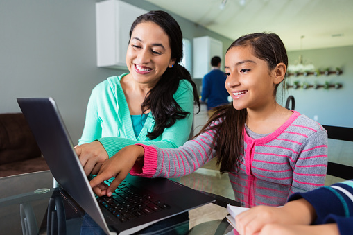 Pretty Hispanic mother and pre-teen daughter point to something on the laptop computer's screen. They smile as they discuss the girl's homework assignment. They are sitting at their glass top kitchen table. The girl's father is in the kitchen in the background.