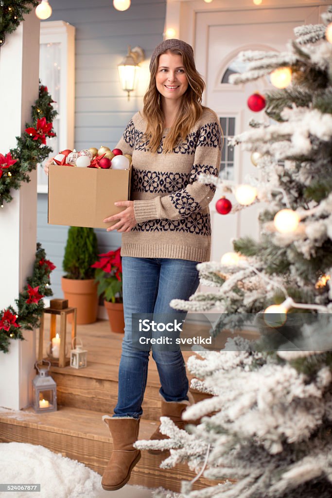 Woman in front of a house. Woman decorating the yard in front of her house. Coming from the house towards tree and holding box with christmas decorations. Wearing knitted sweater, hat and boots. House, yard are decorated with festive string lights, christmas flowers. Day time, winter. Christmas Stock Photo