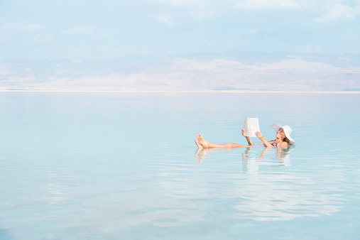 Woman with sun hat holding and reading a newspaper while lying on back with crossed feet, floating in salty water of Dead Sea, Israel, Middle East.