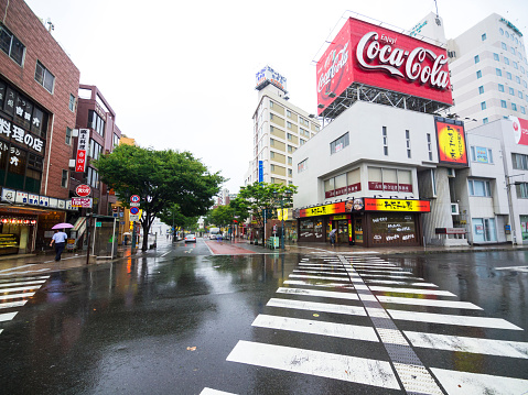 Beppu, Japan - August 9, 2014: Rain day view of street crossing in front of train station in Beppu. There are big crossing lines and buildings in background with lot of commercial panels like Coca Cola one. Above is grey sky.