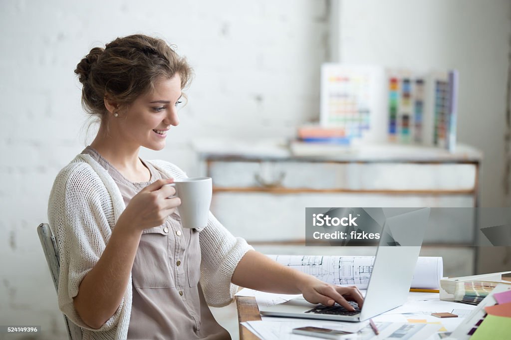 Work with enjoyment Portrait of beautiful happy smiling young designer woman sitting at home office desk with cup of coffee, working on laptop in loft interior. Attractive cheerful model using computer, typing. Indoors Wellbeing Stock Photo