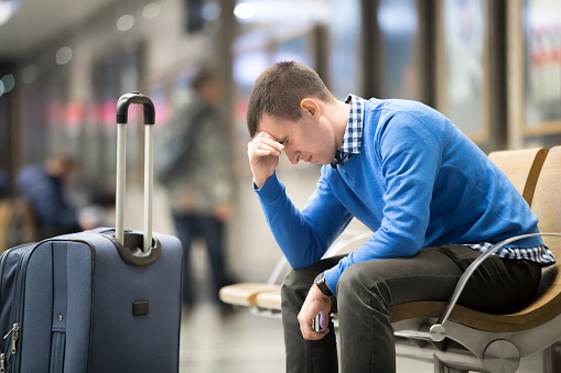 Portrait of young handsome guy wearing casual style clothes waiting for transport. Tired traveler man travelling with suitcase sitting with frustrated facial expression on a chair in modern station