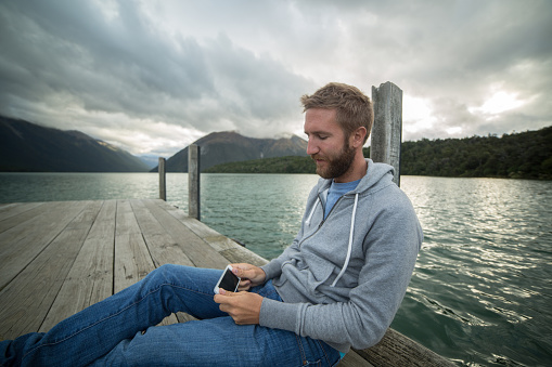 Young man on a lake pier uses a mobile phone. Shot in Tasman region on the South Island of New Zealand.