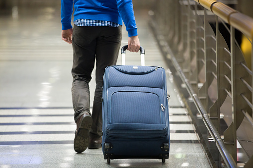 Young man pulling suitcase in modern airport terminal. Travelling guy wearing smart casual style clothes walking away with his luggage while waiting for transport. Rear view. Close-up
