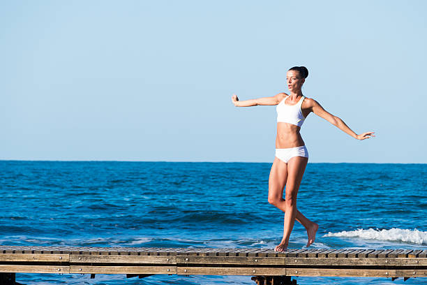 Young woman walking on pier and balcing. Hands outstretched. The sea and the sky on background. Wears white swimsuit.