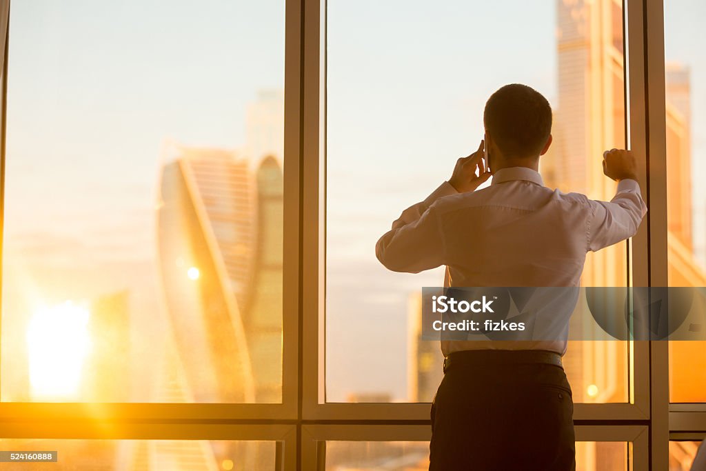 Successful business man Rear view of handsome young businessman standing in confident pose at window in his office and talking on mobile phone while looking at dawn city scenery in panoramic window. Copyspace Rear View Stock Photo