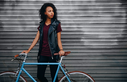 Teenage African American girl looking away seriously while standing on a city sidewalk in front of colourful graffitti with her fixed gear bicycle