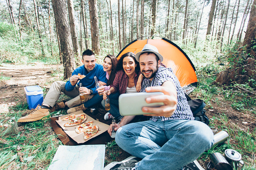Young friends camping and making selfie in front of their tent at the forest. Man sitting and making selfie. All holding bottles with drinks and eating pizza. Cooler on ground.