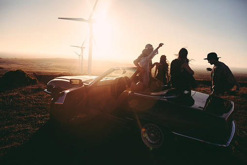 Group of young adult friends is partying at sunset with a guitar and singing leaned to their convertible parked under some large wind turbines