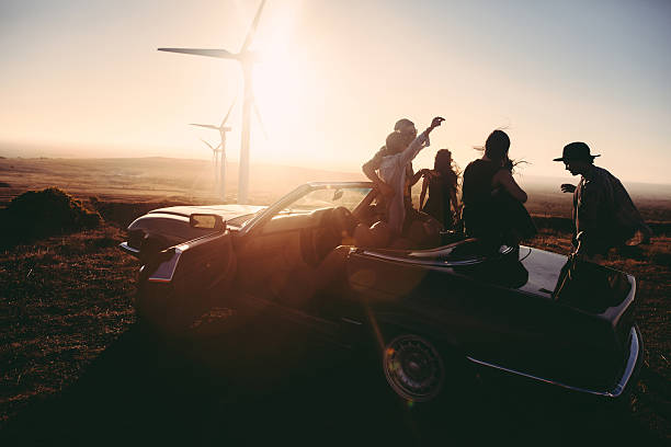Young adult friends enjoing time together with guitar at sunset Group of young adult friends is partying at sunset with a guitar and singing leaned to their convertible parked under some large wind turbines convertible photos stock pictures, royalty-free photos & images