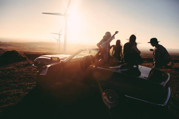 Young adult friends enjoing time together with guitar at sunset Group of young adult friends is partying at sunset with a guitar and singing leaned to their convertible parked under some large wind turbines hipster car stock pictures, royalty-free photos & images