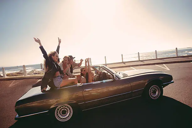 Group of multi-ethnic teenager friends sitting in a convertible celebrating excited for the journey on a summer sunny day