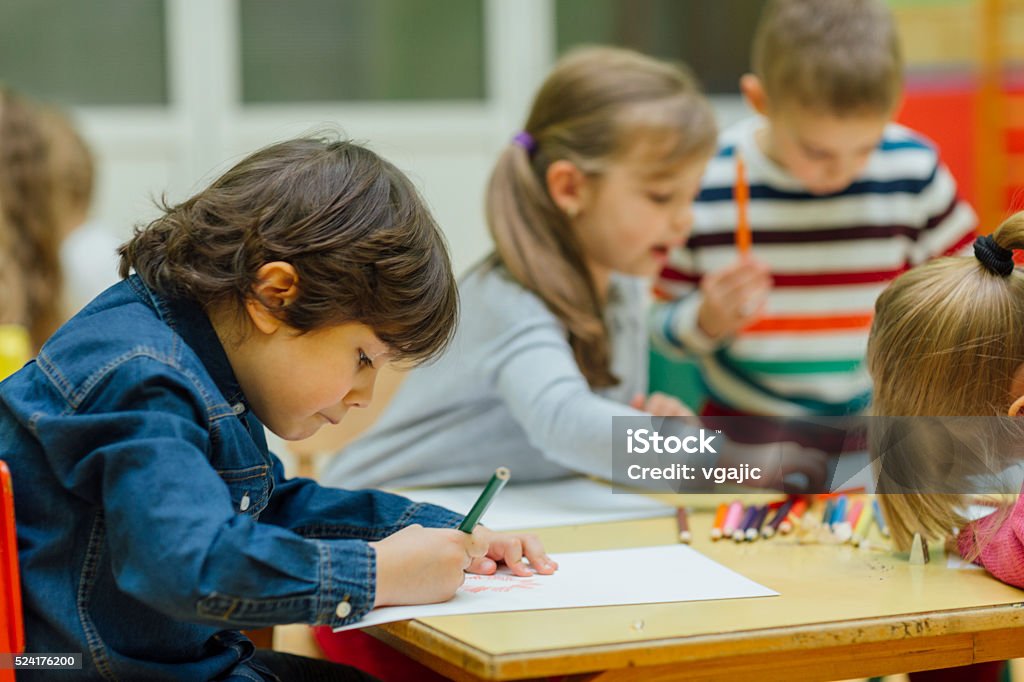 Happy Little Boy In Kindergarten Group of children in kindergarten. Showing their imagination and skill and creativity. Focus to little boy sitting, holding pencil in his hand and drawing. Kids around are talking and drawing. Drawing - Activity Stock Photo