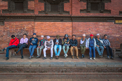 Kathmandu, Nepal - 6th November 2014: Line of local adult men sitting and relaxing on a wooden bench in the historic courtyards of Patan Durbar Square, the UNESCO World Heritage Site in the heart of Kathmandu, Nepal's vibrant capital city.