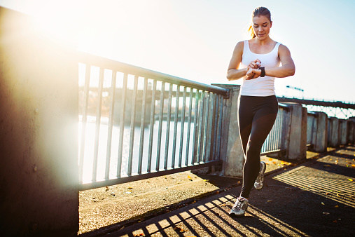 A young athletic woman goes for a morning run, running along the paths at Tom McCall Waterfront Park in Portland, Oregon.  She checks her smart watch, which acts as a pedometer, stopwatch, and heart rate monitor.  The sun shines from behind the Burnside bridge, giving a warm glow to the image.  Horizontal with copy space.